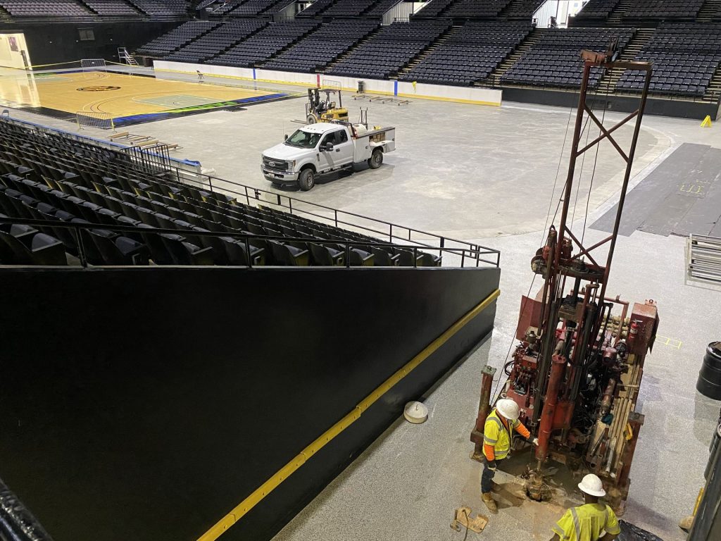 Wide view of geotechnical drilling setup inside arena showing equipment and site conditions during subsurface testing